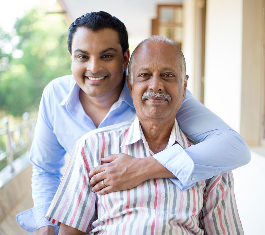 An older man with his arm around his seated father, posed for a photo. They're smiling.